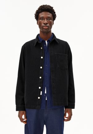 Veste en velours côtelé noir avec un design à col, dotée de boutons pression blancs, d'une poche poitrine, portée par-dessus une chemise bleue à boutons.