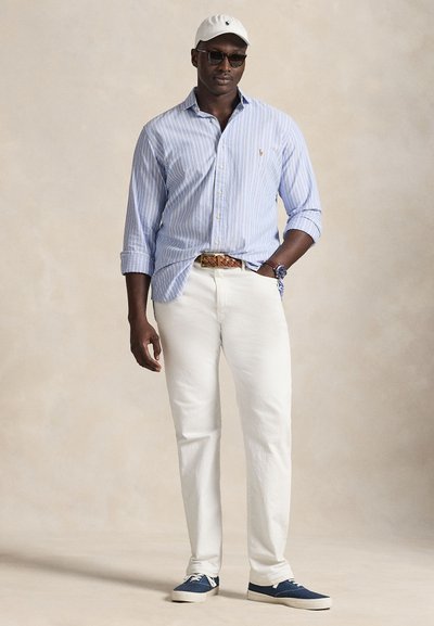 Vita bomullbyxor, ljusblå randig skjorta, vitt keps, solglasögon och blå sneakers. Märkbar bältesdetalj synlig.