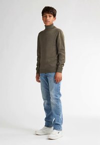 Zielony golf w kolorze oliwkowym, dopasowany fason, prążkowane mankiety, zestawiony z jasnoniebieskimi dżinsami i białymi butami sportowymi. Prosty, casualowy wygląd.