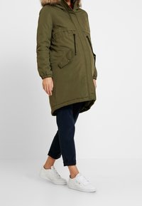 Person i en olivgrön graviditetparkas med pälsbeklädd huva, marinblå uppvikta byxor och vita sneakers, som står framför en vit bakgrund.