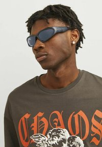 Schwarze Sportsonnenbrille mit schlankem Design und dunklen Gläsern. Getragen mit einem grauen T-Shirt, das großen orangefarbenen Text und einen grafischen Druck zeigt.