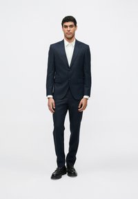 Tengerészkék öltöny, testhezálló zakóval és egyenes szárú nadrággal, világos színű inggel és fekete cipővel, fehér háttér előtt állva.