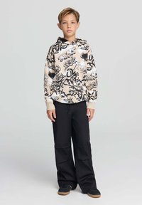 Felpa beige con stampe in stile graffiti nere, abbinata a pantaloni neri a gamba larga e sneakers scure, caratterizzata da una vestibilità rilassata e coulisse.