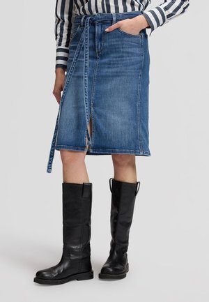 Personne portant une jupe en jean bleu longueur genoux avec une fente à l'avant et une ceinture à nouer, des bottes noires montantes et une chemise rayée noire et blanche.