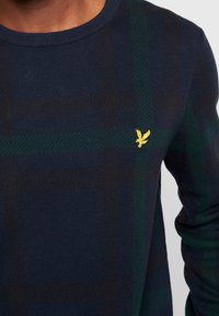 Maglione navy con motivo a quadri in verde scuro e nero, caratterizzato da un piccolo logo ricamato giallo sul petto e un colletto girocollo.