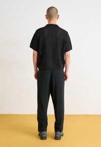Black short-sleeved knitted top with open weave pattern, paired with loose black trousers and gray footwear. Neutral background and flooring.