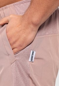 Ljust rosa shorts med en strukturerad yta, som har en sidficka och en synlig märkestagg som heter "SMILODOX." Slät tygfinish.