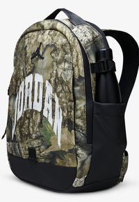 Batoh s maskáčovým vzorom a čiernymi akcentmi, obsahujúci výrazné biele logo „Jordan“, bočnú kapsu na fľašu s vodou a polstrované popruhy.