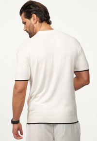 Kurzarm-Polohemd in Creme mit strukturiertem Finish. Besondere Merkmale sind schwarze Besätze an den Ärmeln und dem Saum. Entspannt geschnitten mit einem klassischen Kragendesign.