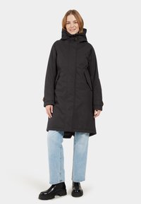 Schwarze wasserdichte Parka mit Kapuze, Reißverschluss, Seitentaschen und geknöpften Manschetten, kombiniert mit hellblauen Jeans und schwarzen Stiefeletten.