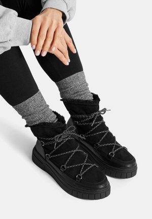 Ręce spoczywają na kolanach, nosząc czarne legginsy, szare skarpetki z dzianiny oraz czarne zimowe buty z grubymi podeszwami i sznurowadłami w kształcie krzyża.