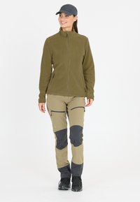 Fleecejacka i olivgrön, med dragkedja, kombinerad med khakibyxor som har svarta lappar och fickor med dragkedja. Svarta vandringskängor.