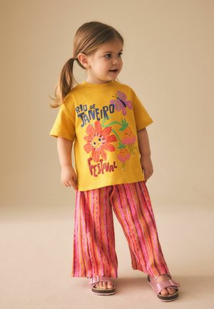 Fată tânără cu păr castaniu prins în coadă de cal, îmbrăcată cu un tricou galben „Festivalul Rio de Janeiro“, pantaloni roz cu dungi și sandale roz, în picioare.