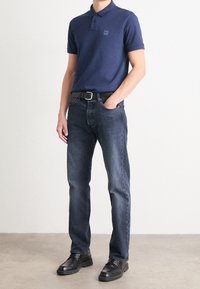 Námořnická polo košile s třemi knoflíky a nášivkou loga, doplněná tmavě modrými džíny a černými botami. Jednoduché pozadí, minimální detaily.