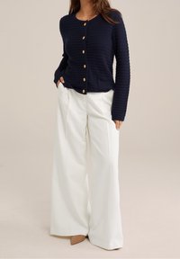 Granatowy prążkowany sweter rozpinany z okrągłym dekoltem, drewnianymi guzikami i przednimi kieszeniami, zestawiony z białymi szerokimi spodniami i beżowymi butami na obcasie.