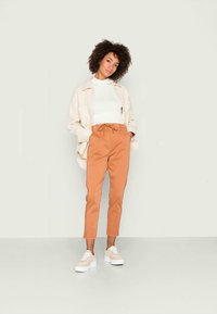 Calças de algodão bege com cintura elástica, combinadas com uma blusa branca ajustada e uma jaqueta oversized bege. Ténis brancos de plataforma completam o visual.