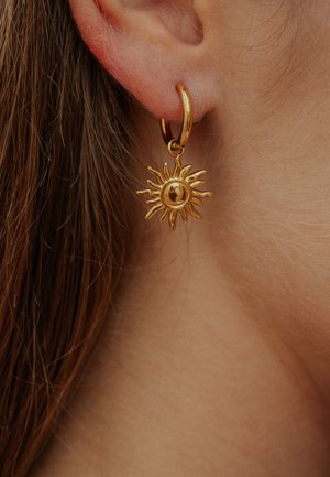 Cercei aurii tip cerc cu un design de raze solare ascuțite și un detaliu rotund central, atârnând aproape de lobul urechii.