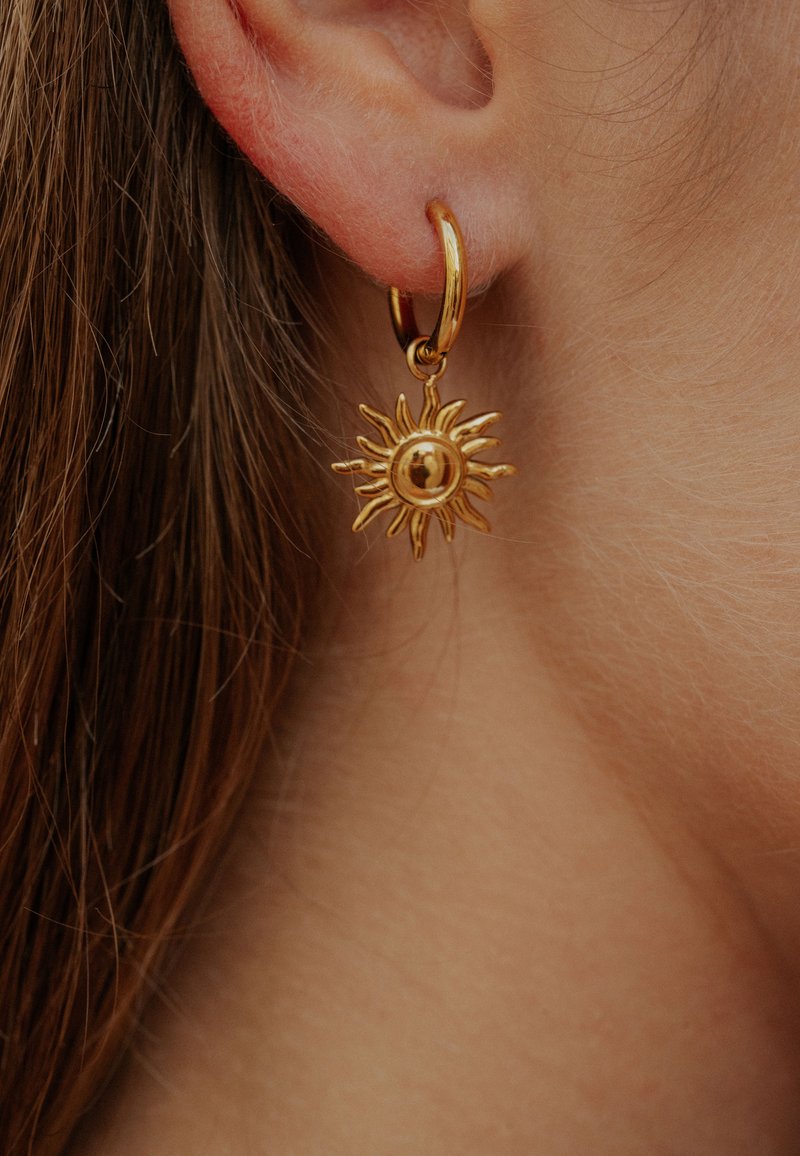 Arany karika fülbevalók, napkitörést formázó mintával, hegyes sugarakkal és központi, kerek részlettel, közel lógva a fülcimpa mellett.