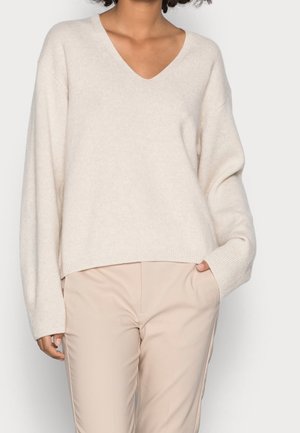 Personne portant un pull col en V crème oversize et un pantalon beige, avec une main dans la poche, sur fond uni.