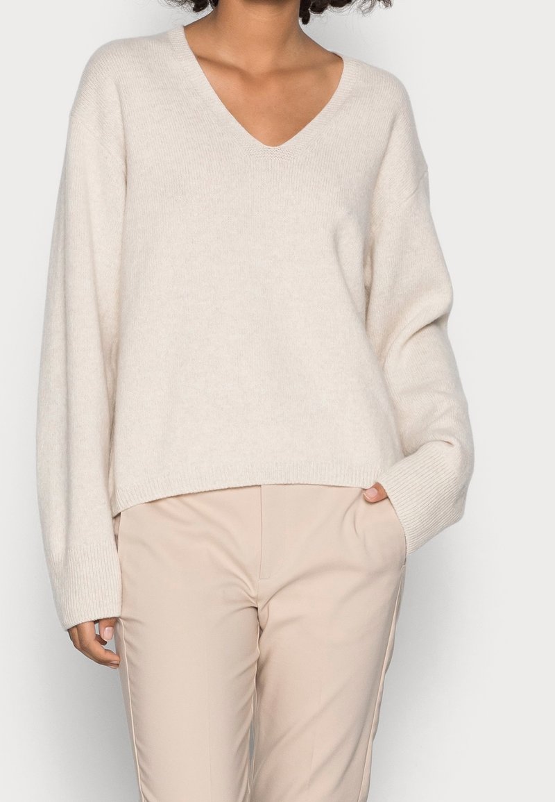 Personne portant un pull col en V crème oversize et un pantalon beige, avec une main dans la poche, sur fond uni.