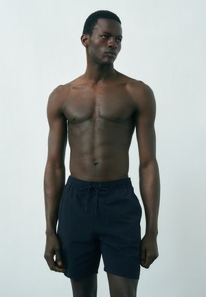 Hemdloser Mann mit dunkler Haut, der marineblaue Badehose mit Kordelzug trägt und vor einem einfarbigen hellgrauen Hintergrund steht.