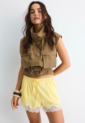 Frau trägt ärmellose braune cropped Jacke mit hohem Kragen und gelbe Shorts mit Kordelzug und weißem Spitzenbesatz, posiert mit Armbändern an einem Arm.