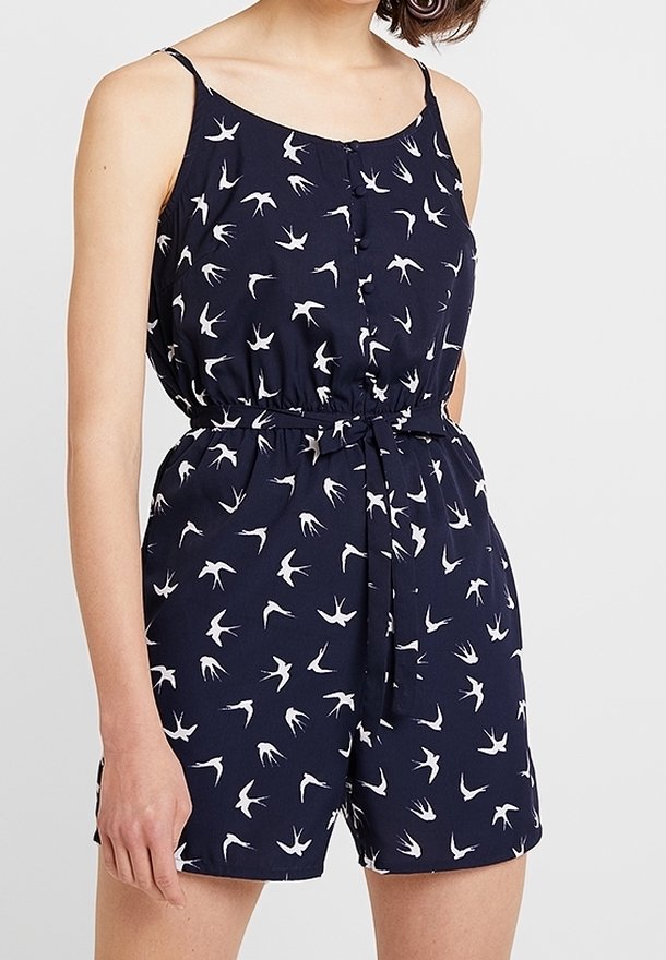 Femme portant une combinaison sans manches bleu marine avec des motifs d'oiseaux blancs et une ceinture nouée à la taille.