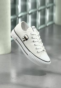 White sneaker with black trim, lace-up design, and a small cartoon figure emblem on the side, displayed against a blurred background.