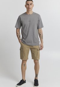 T-shirt en coton gris à manches courtes, avec un petit logo. Short cargo beige avec poches latérales et coupe décontractée. Baskets noires.
