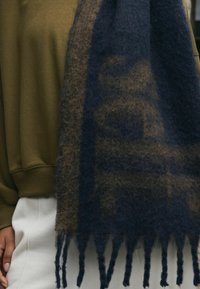 Granatowo-brązowy szalik w kratę wykonany z miękkiego, puszystego materiału, z frędzlami na krawędziach i prostokątnym kształcie, ułożony na zielonej bluzce.
