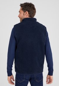 Marineblaues Fleece-Weste aus strukturiertem, weichem Material, mit hohem Kragen und ärmellosem Design. Über einem langärmeligen Hemd getragen, mit lässiger Passform.