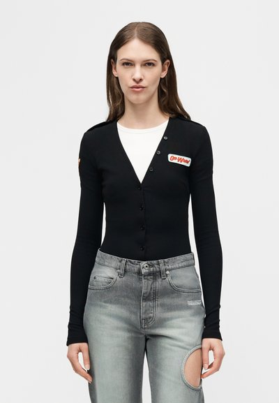Jeune femme portant un cardigan noir boutonné sur un haut blanc et un jean gris avec une grande découpe circulaire sur une cuisse.
