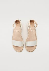 Sandales espadrilles beiges avec une large sangle blanche brillante, semelle intérieure beige et bords tressés, ornées de clous métalliques sur les sangles de talon.