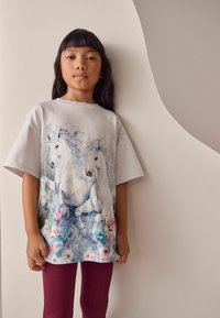Weißes übergroßes T-Shirt mit einem Einhorn-Design in Blau- und Rosatönen, kombiniert mit gerippten kastanienbraunen Leggings. Sanfte Baumwollstruktur.