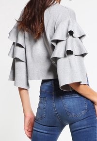 Grau geschnittener Pullover mit geschichteten Rüschärmeln, aus einem weichen Material. Kombiniert mit blauen, figurbetonten Jeans.