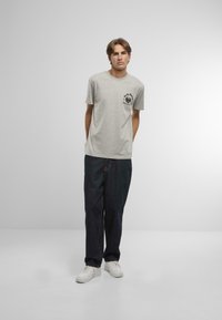 Tricou gri cu mânecă scurtă, cu un grafic negru pe buzunarul din stânga pieptului, asortat cu blugi bleumarin și adidași albi.