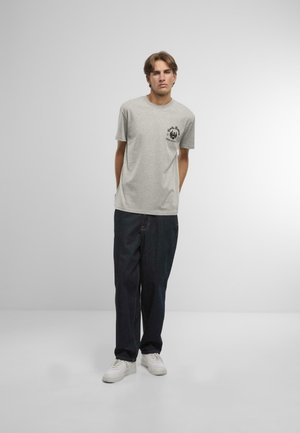 Šedé tričko s krátkým rukávem a černou grafikou na levé kapse, kombinované s tmavě modrými džíny a bílými teniskami.