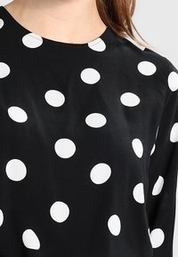 Haut en tissu noir orné de gros pois blancs, avec un col rond et des manches longues, texture lisse avec un léger éclat.