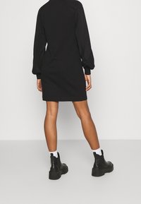 Osoba oblečená v černých dlouhých šatech, bílých ponožkách a černých kotníkových botách, stojící zády k fotoaparátu na jednobarevném pozadí.