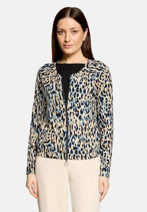 Kvinde iført en langærmet, leopardmønstret jakke med lynlås over en sort top, kombineret med lyse beige bukser, stående mod en ensfarvet baggrund.