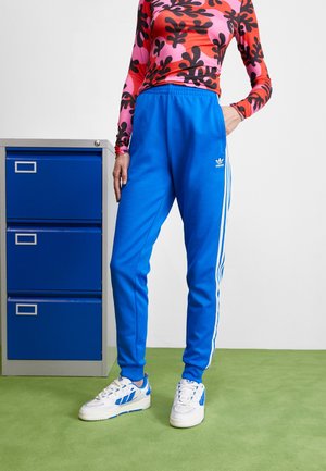 Personne debout sur une moquette verte, portant un pantalon de survêtement bleu Adidas, des baskets blanches Adidas et une chemise à motifs roses à manches longues à côté d'une armoire à dossiers bleue.