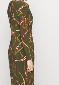 Person klädd i en långärmad olivgrön klänning med ett sammanflätat bälte och kedjemönster i bruna, gyllene, röda och vita nyanser.