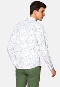 Biała koszula z długim rękawem, w niebieski kwiatowy wzór, z zapięciem przy kołnierzyku i prostym tyłem. Noszona z zielonymi spodniami.