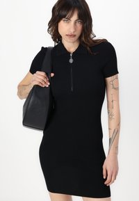 Black ribbed dress with short sleeves and a zippered collar. Accompanied by a black shoulder bag. Minimalist and form-fitting design.