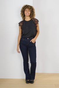 Črna bluzica z čipkastimi rokavi, v kombinaciji s temno modrimi širokimi jeans hlačami. Model nosi ravne črne čevlje in stoji v minimalističnem okolju.
