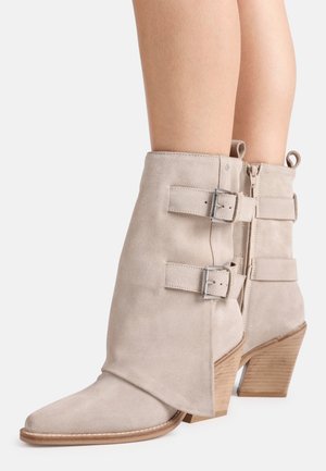 Bottines en daim beige à talons en bois empilés, fermeture éclair latérale et deux sangles réglables avec boucle, portées sur des jambes nues.