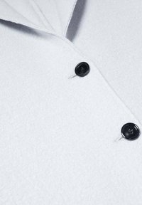 Lichtgrijze, gestructureerde stof close-up van een jas met twee zwarte knopen. Het ontwerp bevat een kraag en zichtbare stiksels.