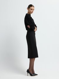 Svart blus med långa ärmar i texturerat tyg, kombinerad med en figurnära, knälång svart kjol. Modellen bär svarta högklackade skor och transparenta strumpbyxor.