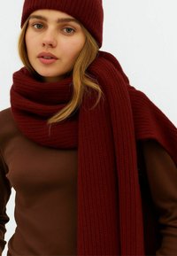 Ung kvinde med lyst hår iført en brun top, en tykk bordeaux strikket halstørklæde viklet om halsen og en matchende strikket hue.