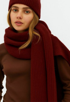 Ung kvinde med lyst hår iført en brun top, en tykk bordeaux strikket halstørklæde viklet om halsen og en matchende strikket hue.